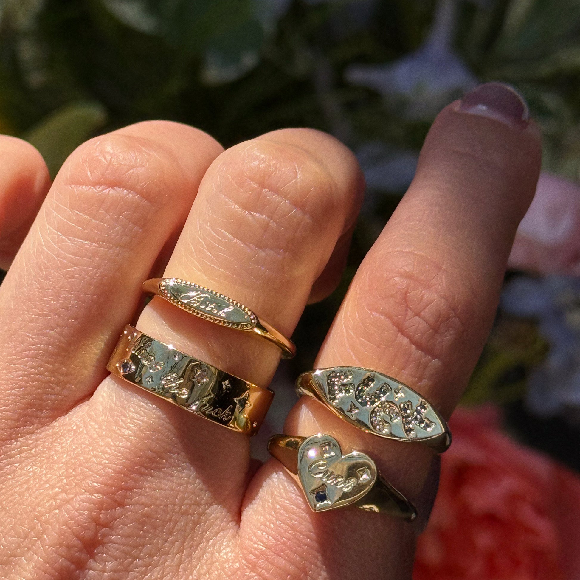 A hand gleams in sunlight, wearing four Colorful Language Fuck Signet Rings in 14k yellow gold—each one uniquely engraved and designed—set against blurred flowers for a bold, colorful statement.