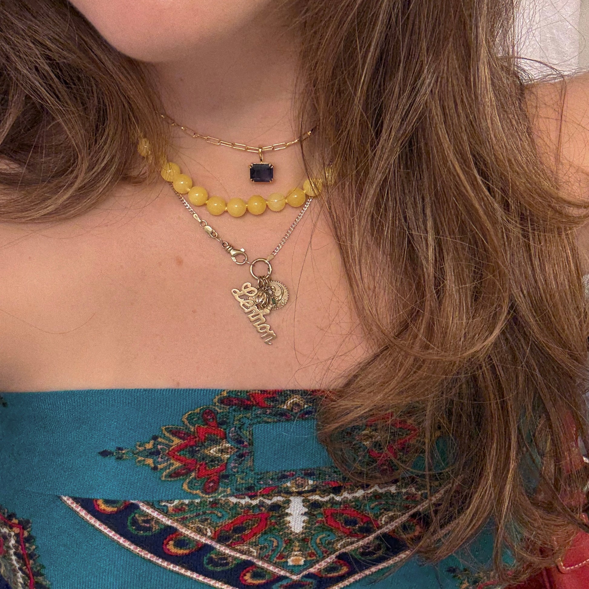 A woman wears layered gold necklaces—a paperclip chain, a yellow bead necklace, the Born to Shine Birthstone Necklace with a dark gemstone pendant, and two more with Lebanon and coin pendants—paired with a teal patterned strapless top.
