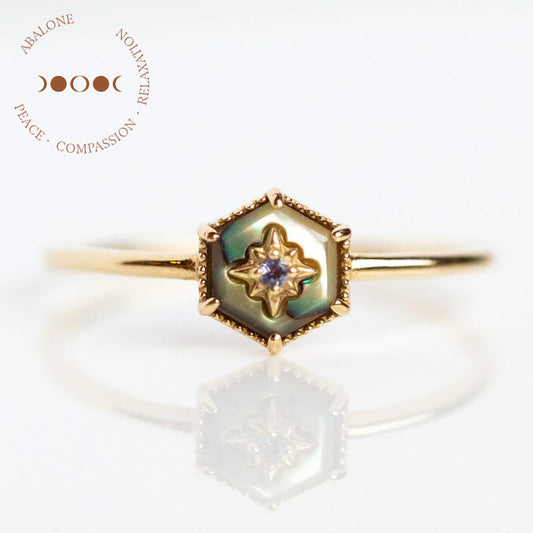 The Solid Gold Crystal Ring for Manifestation features abalone with a hexagonal inlay, an iolite star-set gem, and a four-moon phase symbol. Circular text reads “ABALONE PEACE • COMPASSION • RELAXATION.” Crafted in 9k yellow gold, sample size 7.