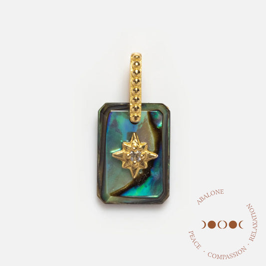 The Solid Gold Crystal Amulet Charm for Manifestation is a rectangular abalone pendant with an iridescent surface, gold bail, starburst design with central gemstone, and Abalone: Peace, Compassion, Relaxation in circular text nearby.