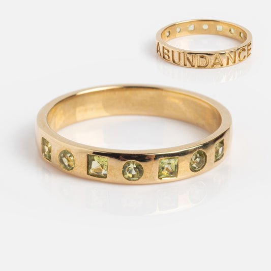 The Solid Gold Within You Birthstone Band (Sample Size 7) features seven natural light green gemstones in a row, set in 9k yellow gold with a 3.1mm band engraved ABUNDANCE inside.