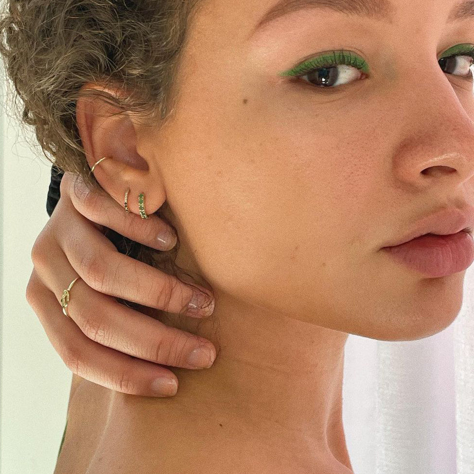 Close-up of a person with curly hair, green eyeliner, and gold hoop earrings wearing the Solid Gold Essential Ear Cuff, gently touching their face. A softly blurred background highlights their striking ear stack.