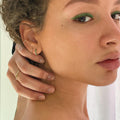 Close-up of a person with curly hair, green eyeliner, and gold hoop earrings wearing the Solid Gold Essential Ear Cuff, gently touching their face. A softly blurred background highlights their striking ear stack.