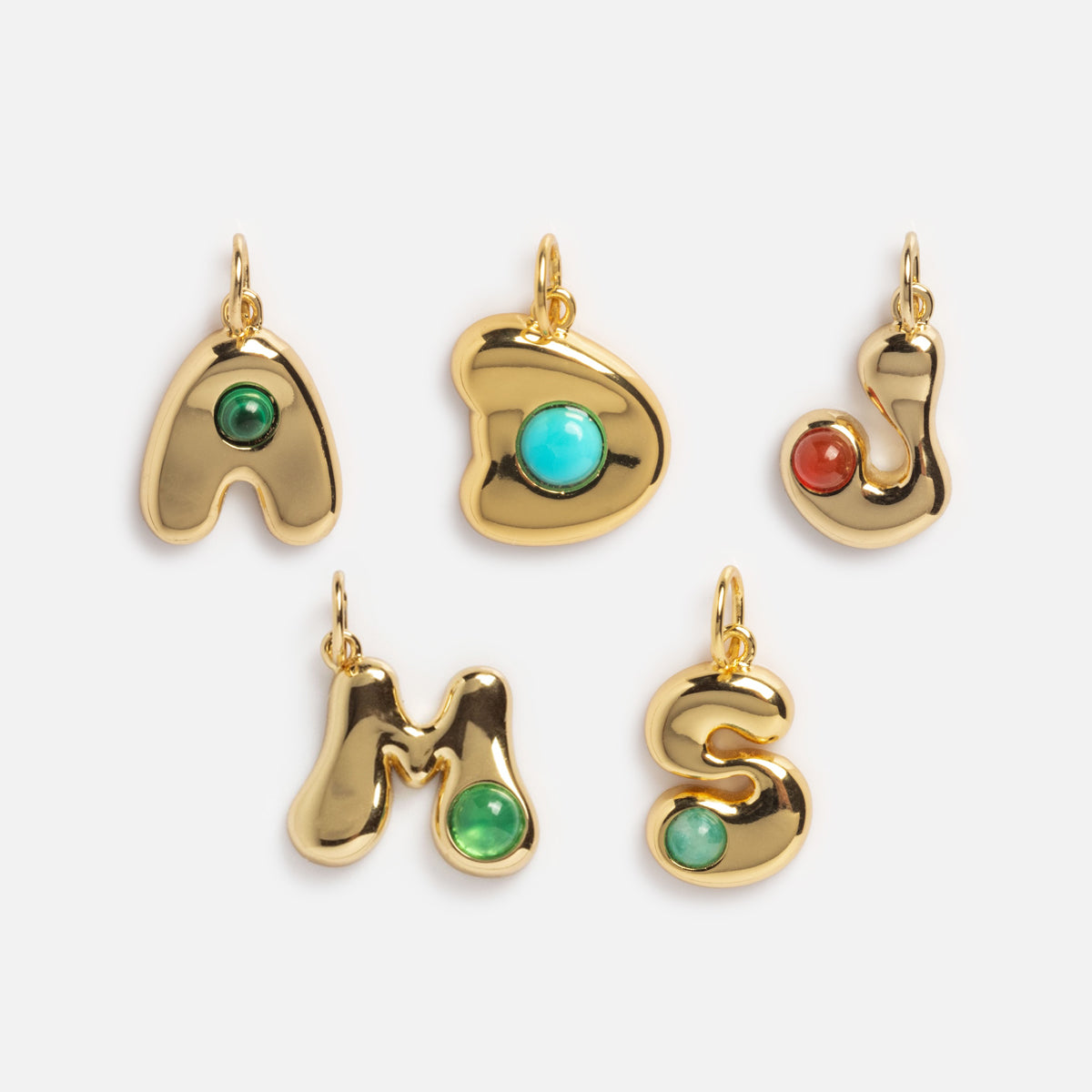Thea Bubble Letter Charm pendants in A, D, J, M, and S feature an 18k yellow gold plated finish with colorful gemstones at the center or side. All five are displayed on a plain white background.
