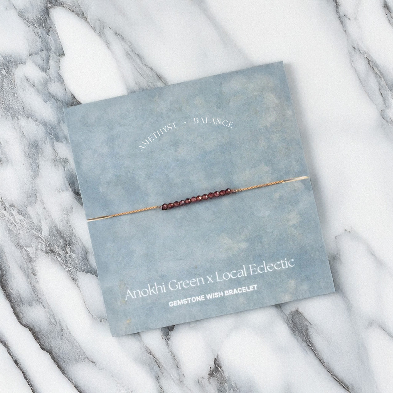 The Anokhi Green x Local Eclectic Gemstone Wish Bracelet, featuring small amethyst beads for balance, is displayed on a blue-gray card labeled with the product name and crafted from natural gemstones.