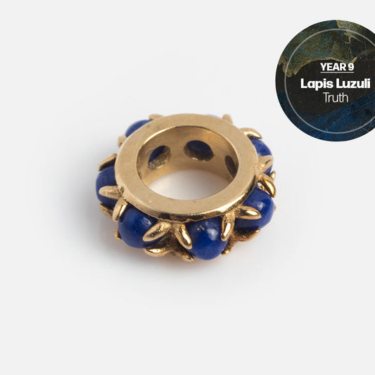 A gold and lapis lazuli bead with gold prongs securing blue stones rests on a white background. In the top right, a circular label reads YEAR 9 Solid Gold Anniversaire Charm—perfect as milestone jewelry.