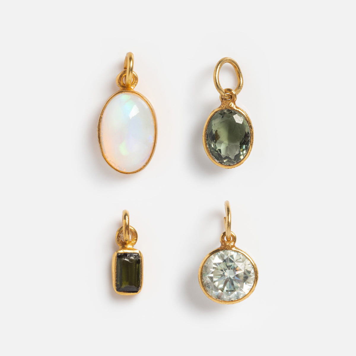 Four small Gemstone Charms by Anokhi Green in 18k gold vermeil—featuring natural oval opalescent, oval green, rectangular dark green, and round clear stones—are arranged on a white background.