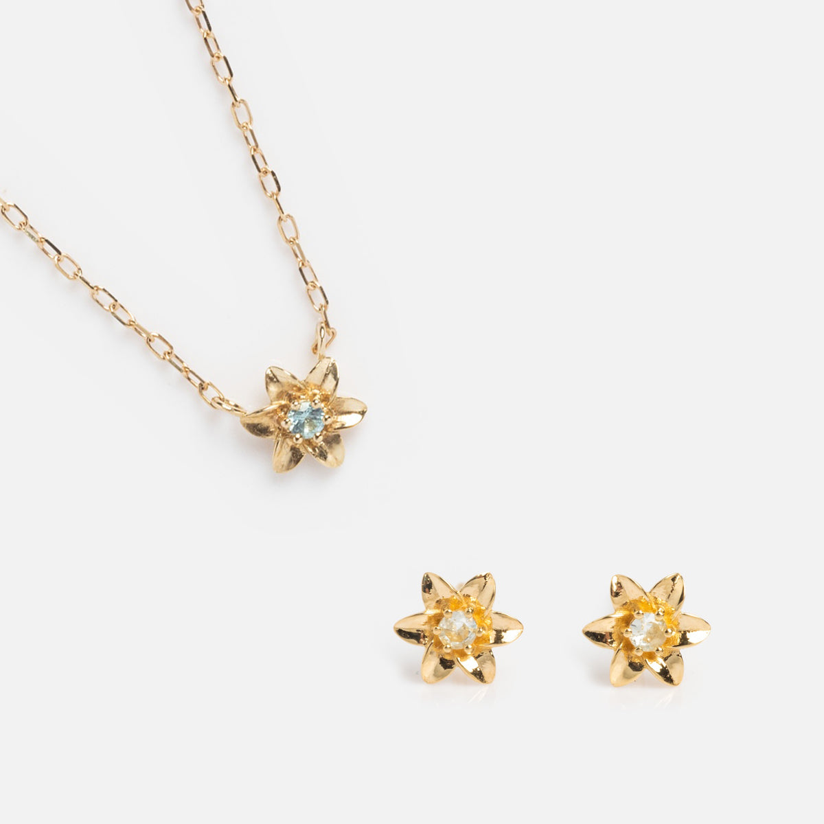 The Solid Gold Birth Flower Gift Set includes a gold necklace with a flower-shaped pendant and matching stud earrings, each with a clear gemstone center—an elegant jewelry gift set shown on a white background.