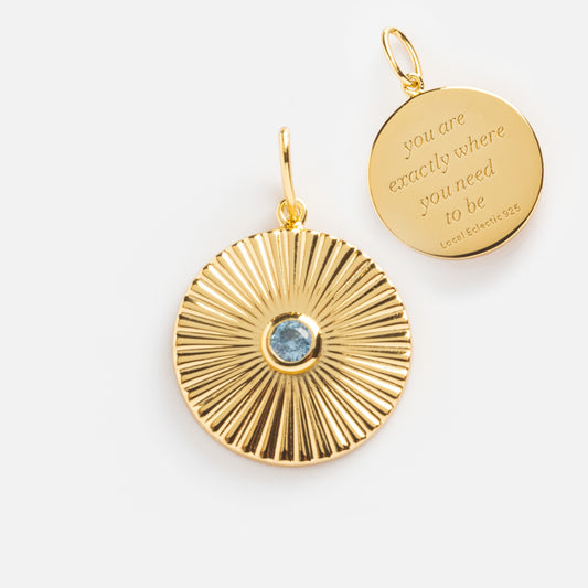 The Mantra Empowerment Coin Charm set features two round 14k gold plated pendants: one with a sunburst and blue gemstone, the other engraved with “you are exactly where you need to be.”.