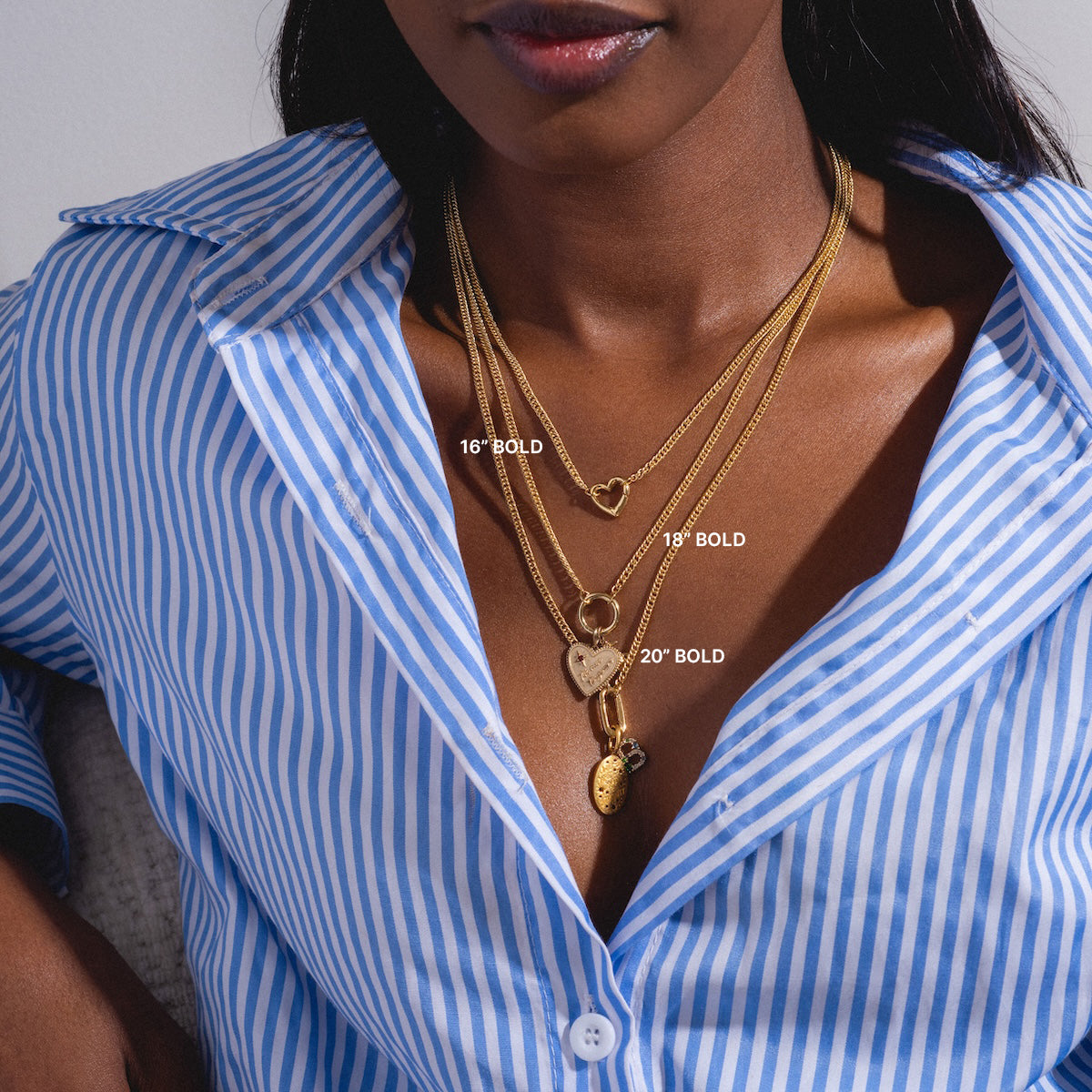 A woman in a blue and white striped shirt wears layered gold-plated necklaces of varying lengths (16, 18, and 20), including the Perfect Start Charm Holder Necklace with unique pendants like a heart on a paperclip chain and an oval charm.