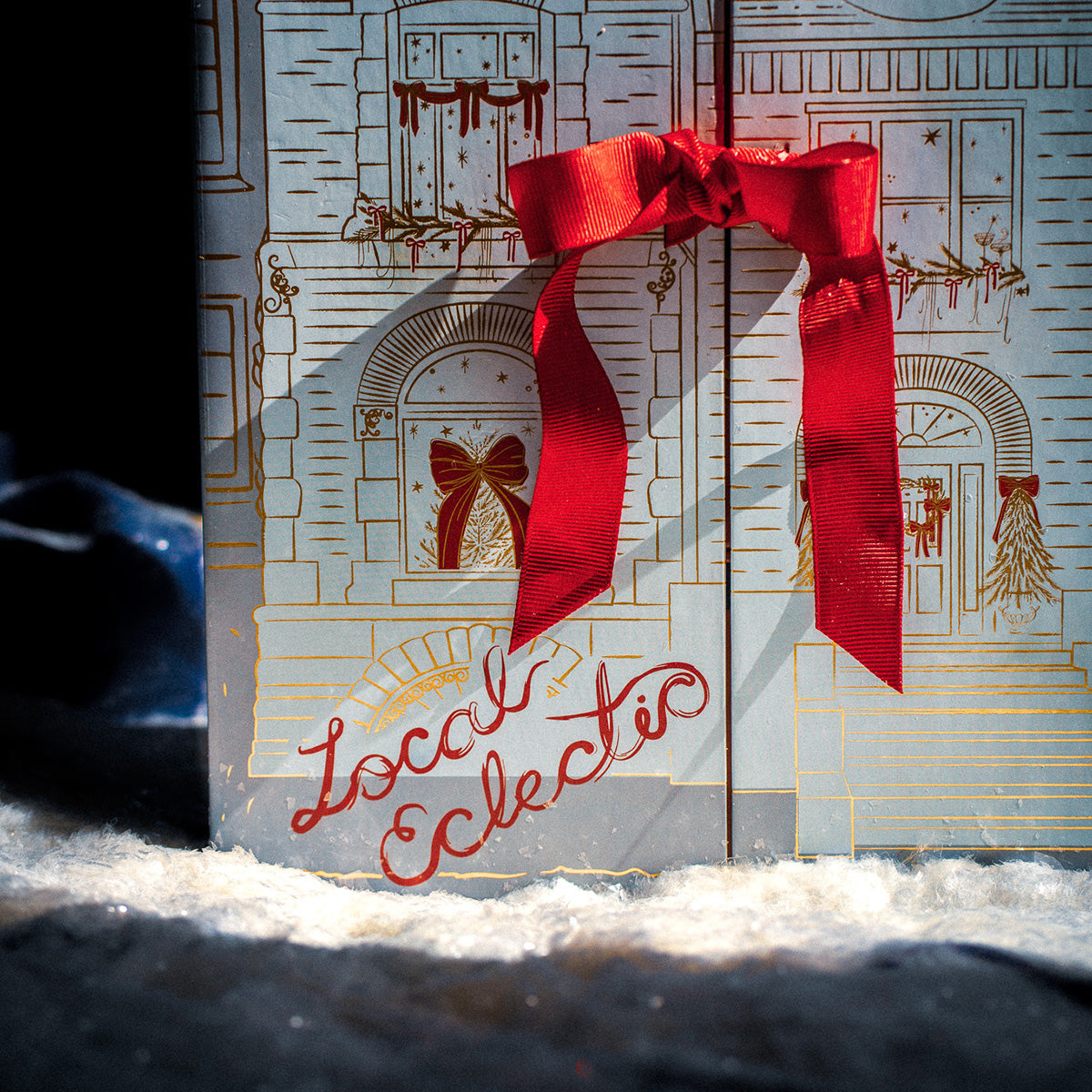A festive 2025 Jewelry Advent Calendar box features an illustrated building, adorned with a red bow and ribbon. “Local Eclectic” is written in red script on the snowy ground in front of the doors.