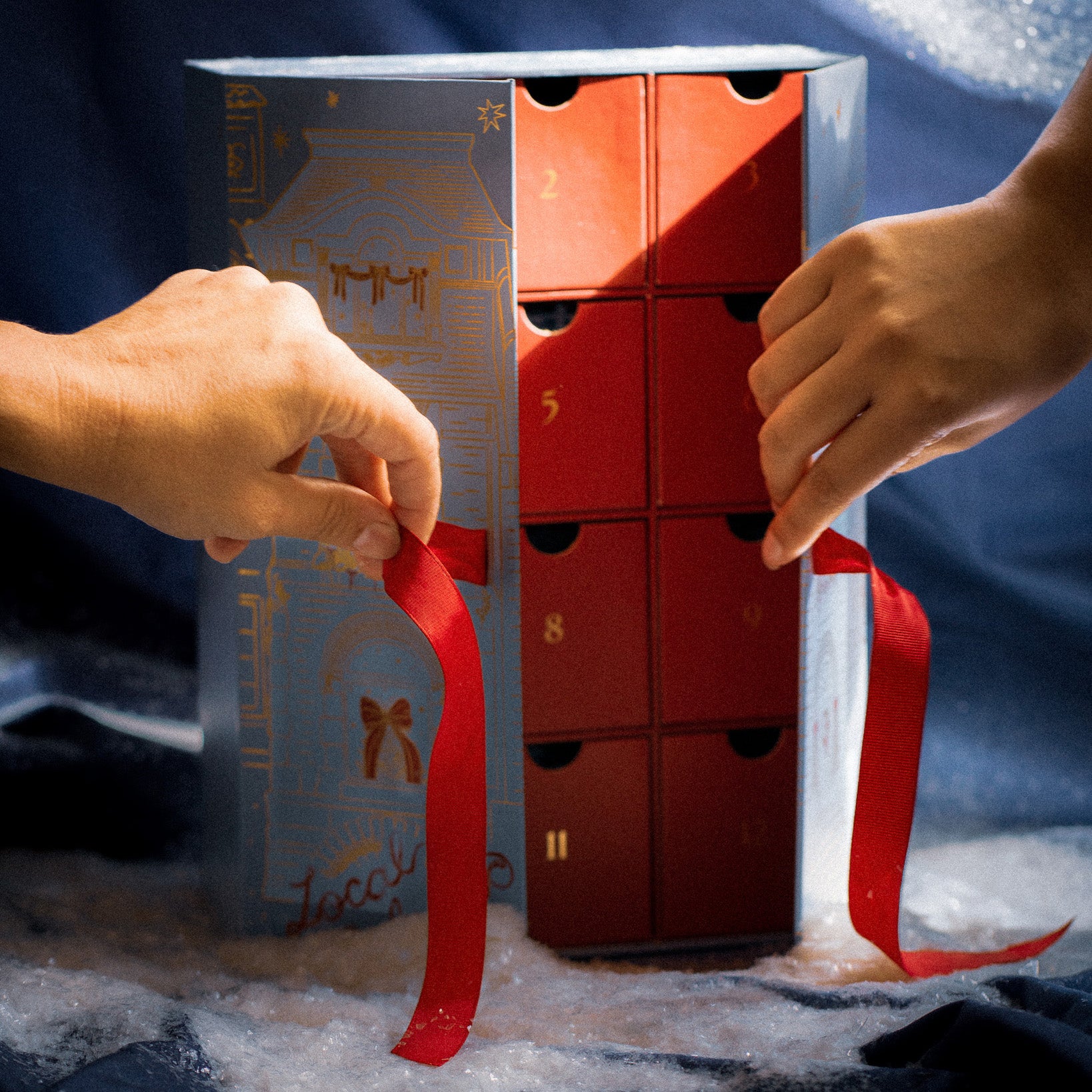 Two hands pull red ribbons on the 2025 Jewelry Advent Calendar, featuring red drawers numbered in gold, set against a snowy, winter-themed background.