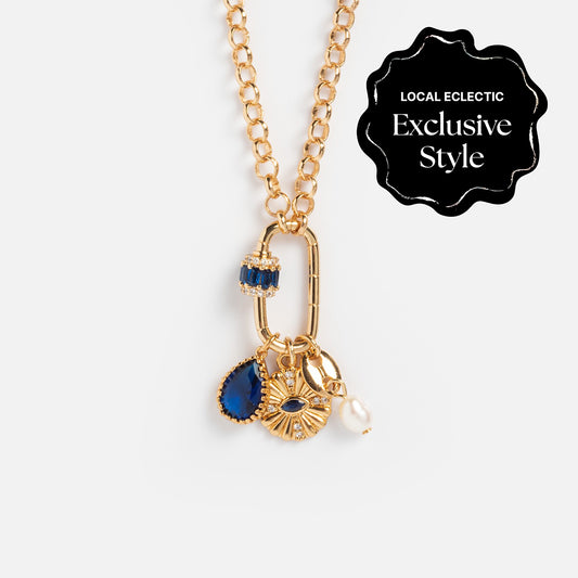 Queen Crystal Birthstone Charm Necklace: Gold adjustable chain with four dangling charms—a blue crystal birthstone, an eye-shaped charm with a blue center, a small pearl, and a ring adorned with blue stones. Exclusive Style label top right.