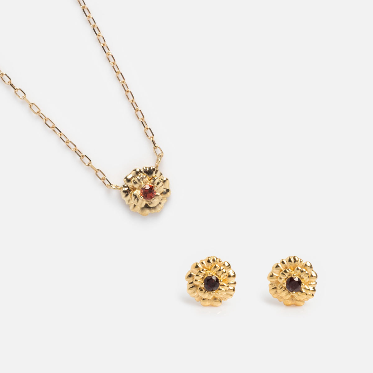 The Solid Gold Birth Flower Gift Set includes a textured circular pendant necklace with a red gemstone and matching gold stud earrings featuring dark gemstones, elegantly displayed on a white background.