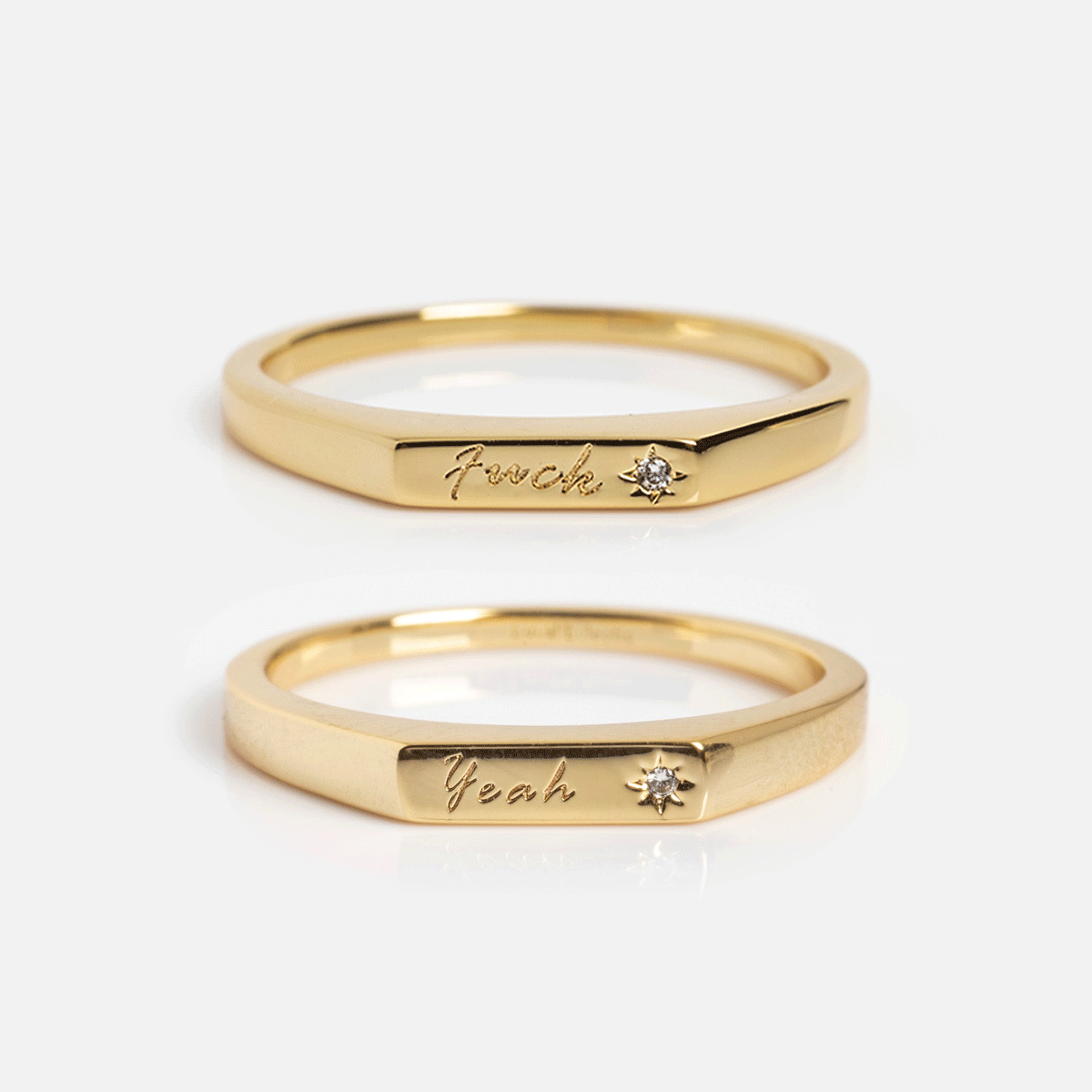 The Tiny Conversation Stacking Rings are two gold bands with flat facets, each engraved in cursive—one reads f*uck, the other yeah—and both have a small diamond accent beside the engraving.