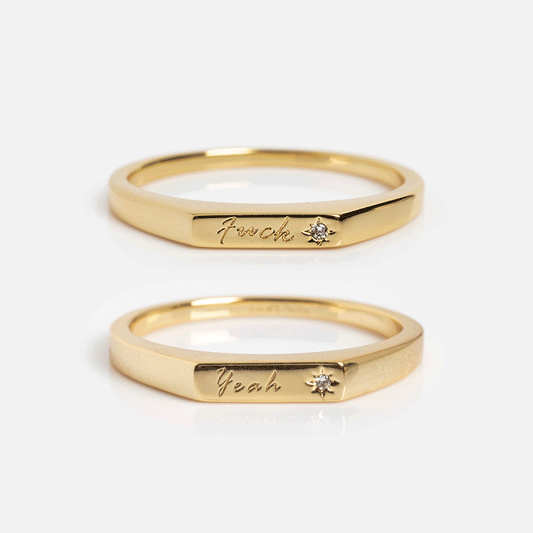 The Tiny Conversation Stacking Rings are two gold bands with flat facets, each engraved in cursive—one reads f*uck, the other yeah—and both have a small diamond accent beside the engraving.