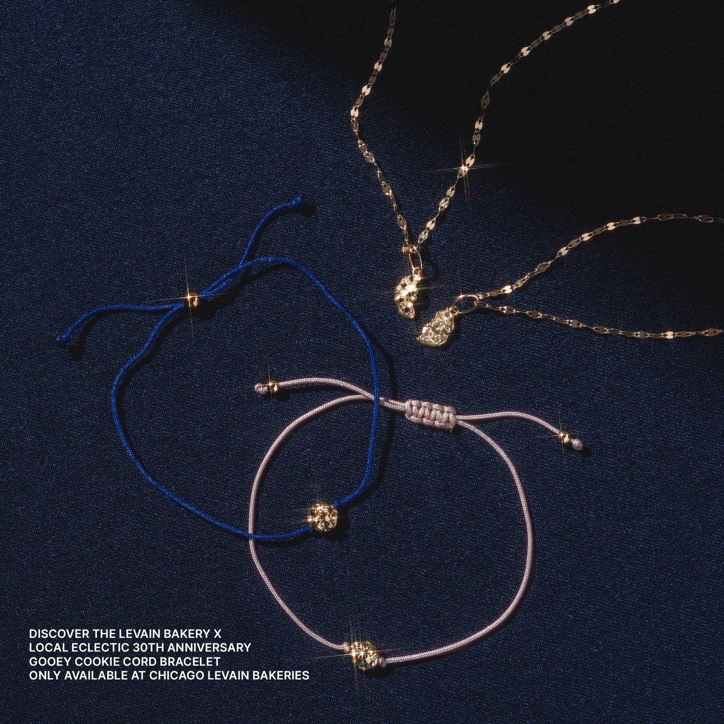 Three jewelry pieces—a gold necklace and two cord bracelets (blue and pink)—are displayed on dark blue fabric. Each features the Levain Bakery x Local Eclectic Solid Gold Share a Cookie Friendship Charm.