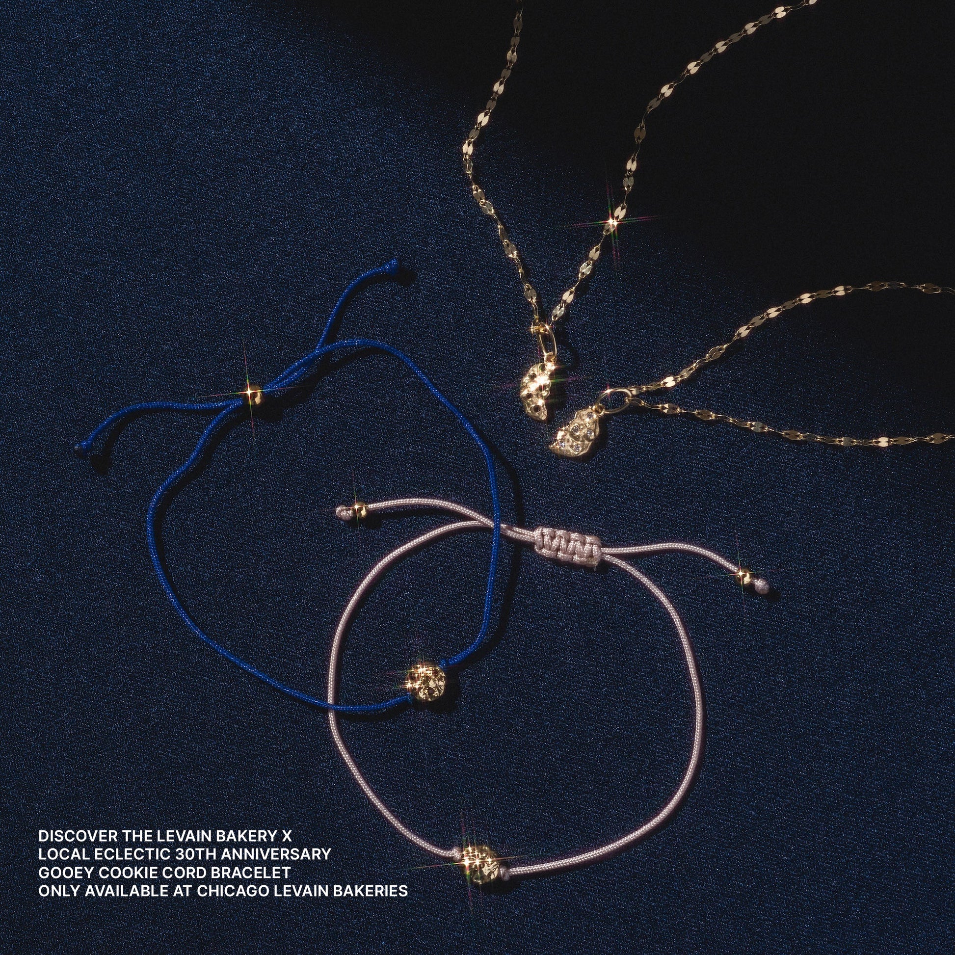 Three jewelry pieces—a gold necklace and two cord bracelets (blue and pink)—are displayed on dark blue fabric. Each features the Levain Bakery x Local Eclectic Solid Gold Share a Cookie Friendship Charm.