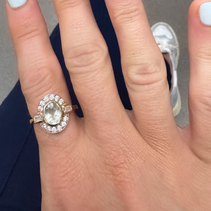 A hand with light blue nails displays the Oval Deco Green Amethyst and CZ Ring, featuring a central green amethyst encircled by petite cubic zirconia on a gold vermeil band. A sneaker and dark pants are visible in the background.
