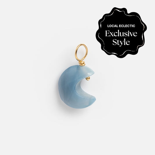 A crescent moon-shaped blue semi-precious stone pendant with a gold loop from the Devi Arts Collective x Local Eclectic Carved Crystal Charms, shown on a white background with a black Local Eclectic Exclusive Style label in the corner.