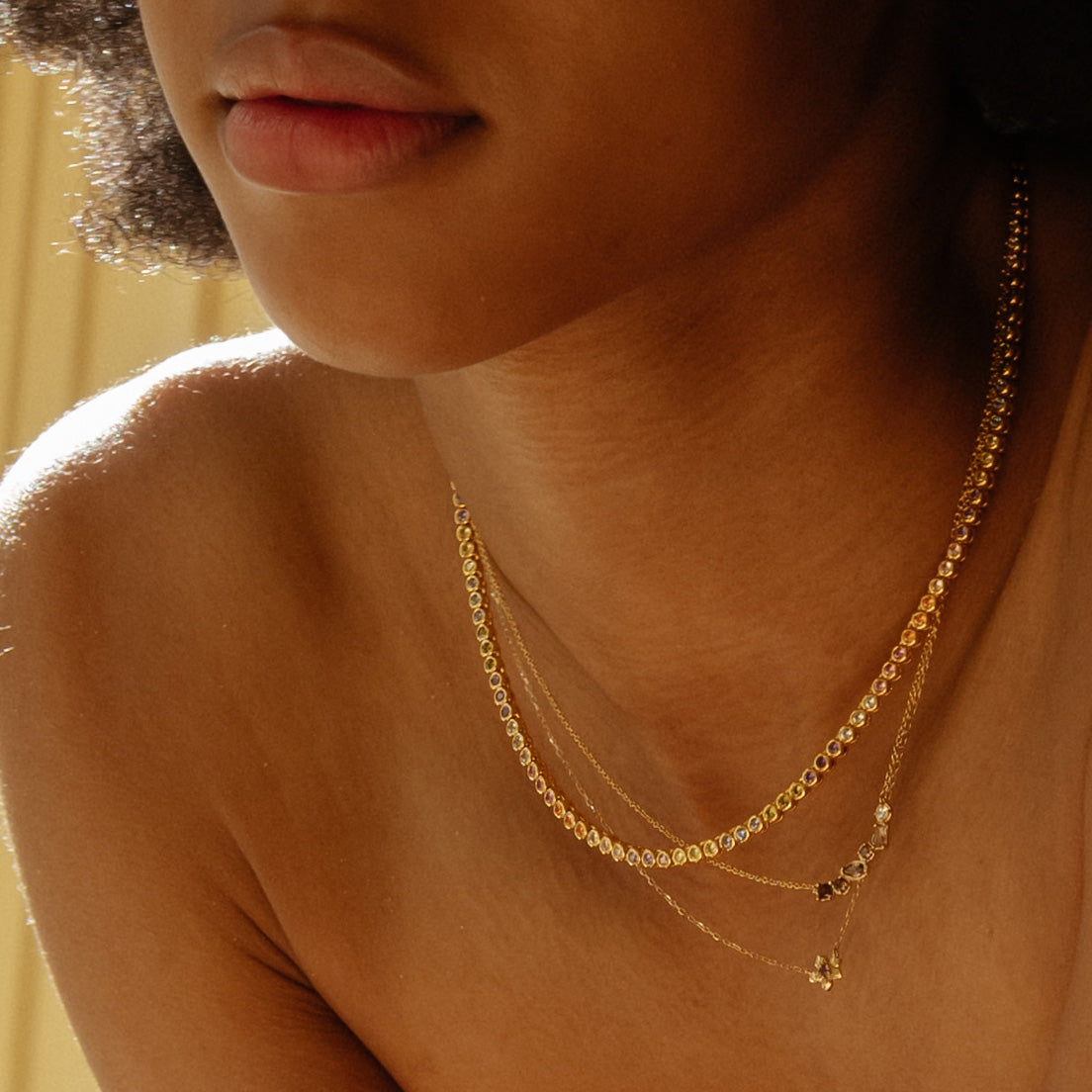 A close-up of a woman’s neck and shoulders showcases the Dreamworks Tennis Necklace, layered with delicate pendants. Soft sunlight highlights her skin and curly hair.