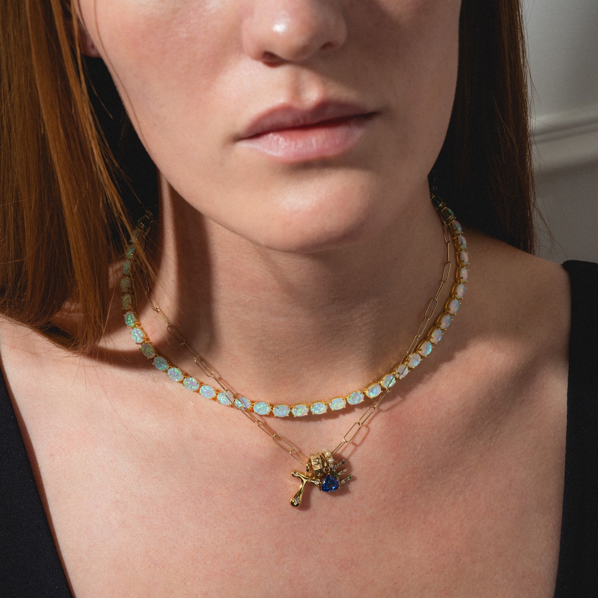 A woman wears two gold necklaces over a black top: one featuring the Star Crossed Celestial Bubble Letter Charm, and another with light blue gemstones. The image is cropped to show her neck and collarbone.