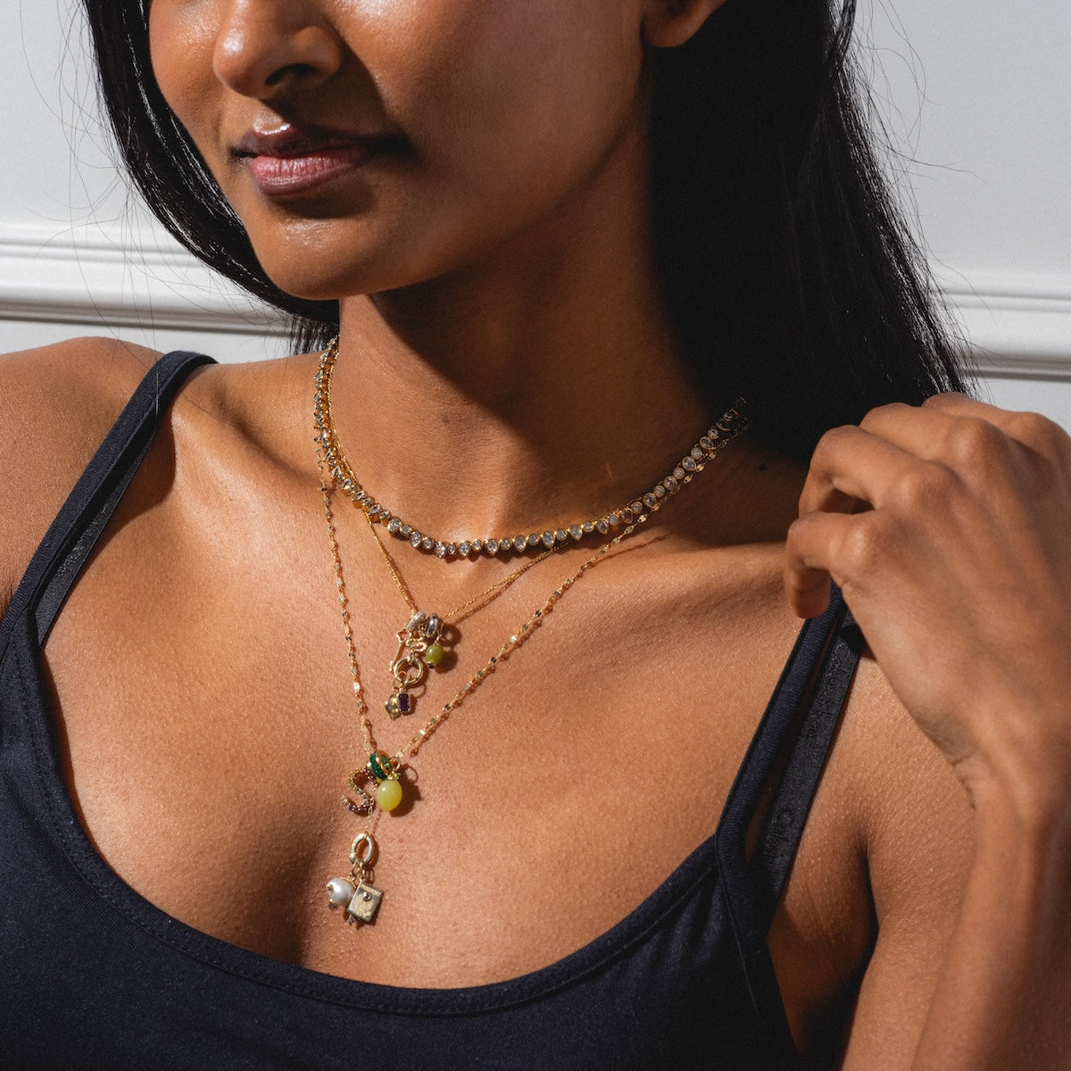 A woman poses against a white background in a black tank top, wearing a necklace stack featuring the Solid Gold Figaro Chain with colorful gemstone pendants.