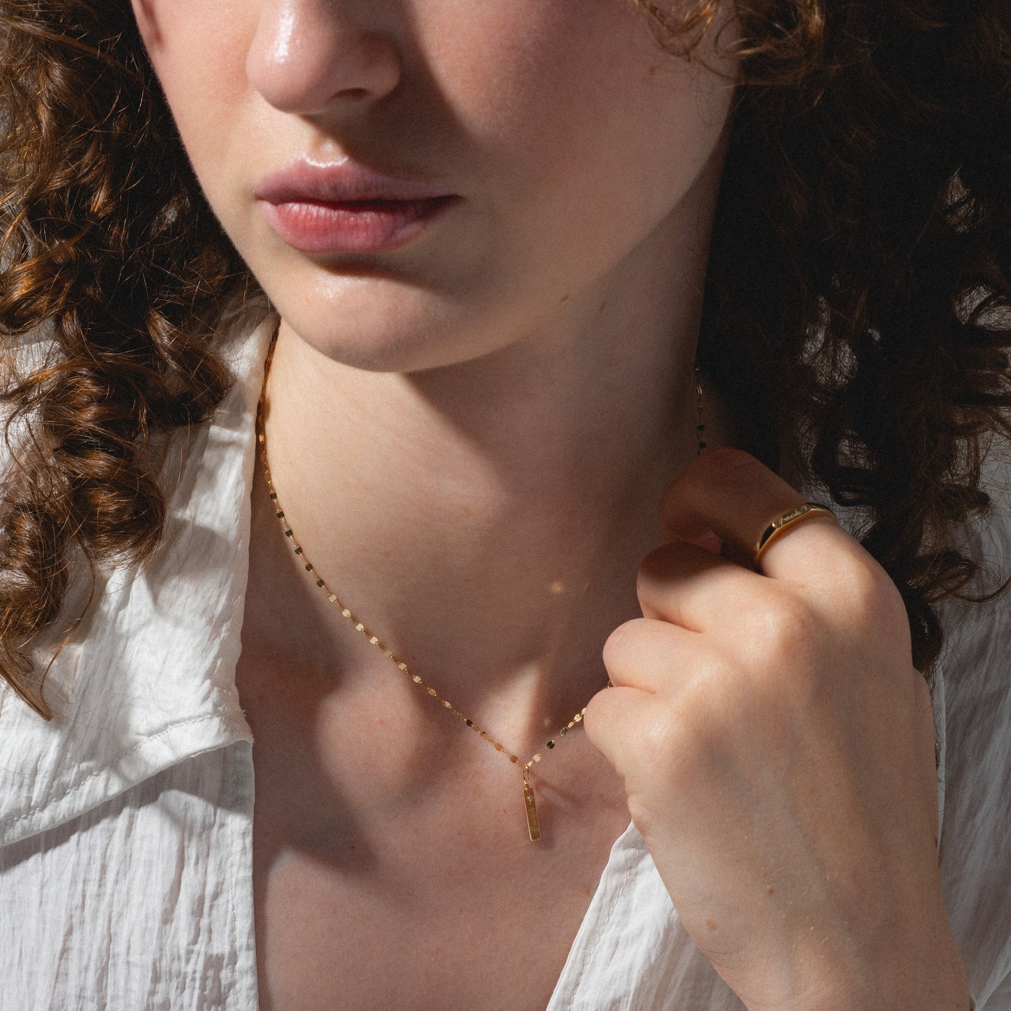 A woman with curly brown hair wears a white shirt, gold ring, and a delicate gold necklace from the Solid Gold Tiny Fuck Gift Set. She holds her Solid Gold Tiny Fuck jewelry and looks slightly away from the camera.