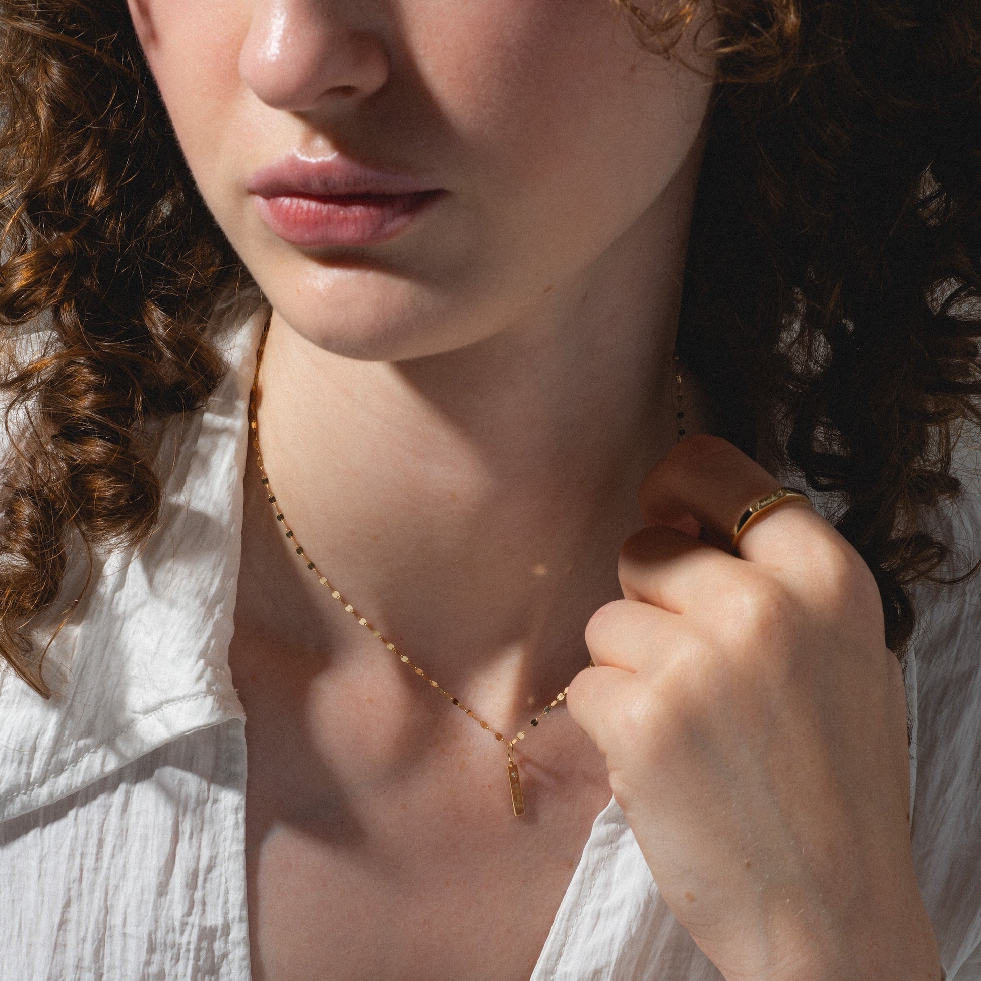 A woman with curly brown hair wears a white shirt, gold ring, and a delicate gold necklace from the Solid Gold Tiny Fuck Gift Set. She holds her Solid Gold Tiny Fuck jewelry and looks slightly away from the camera.