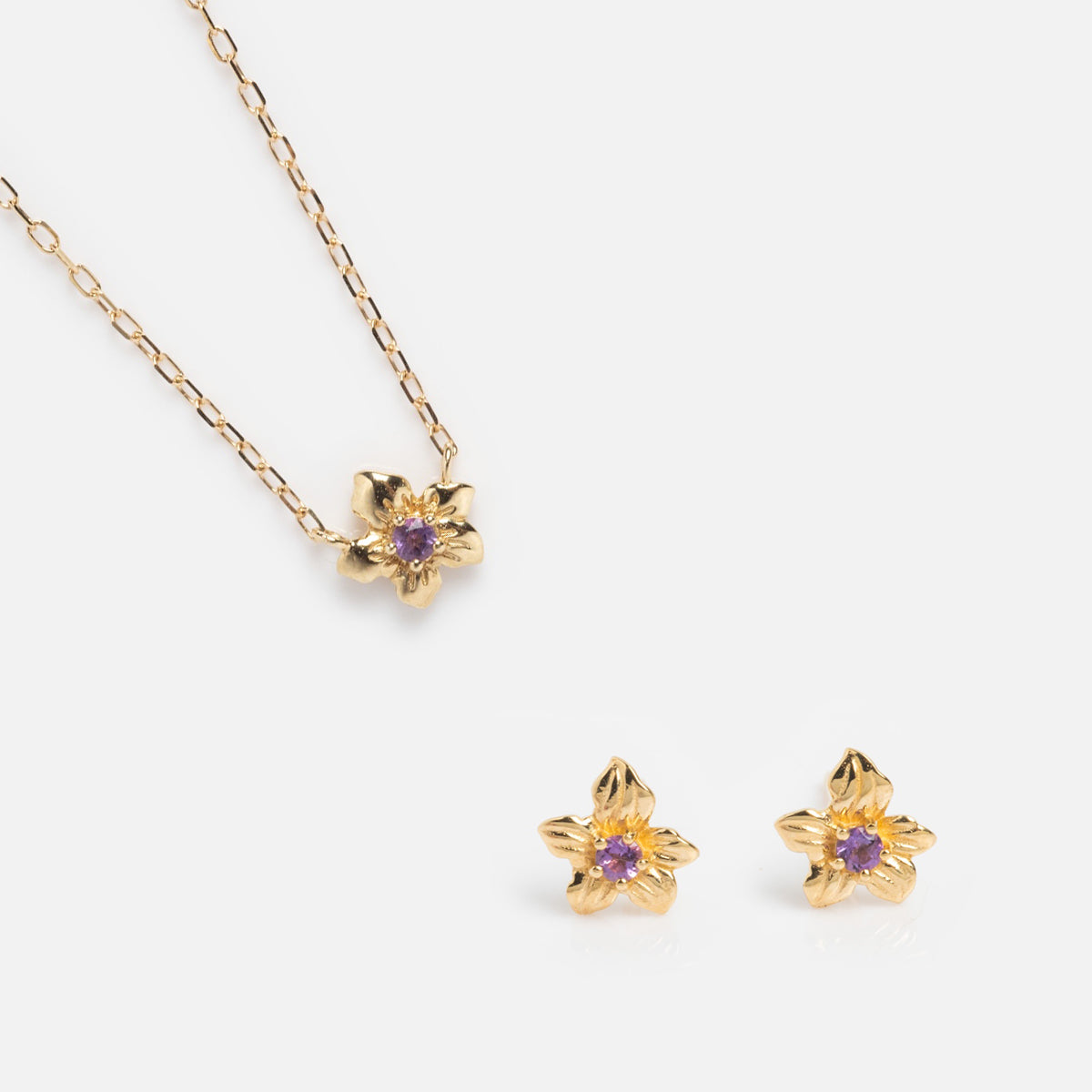 The Solid Gold Birth Flower Gift Set features a gold floral necklace and matching stud earrings, each styled as a five-petal flower with a small central purple gemstone, beautifully displayed on a white background.