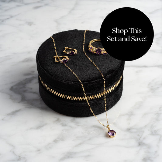 The Solid Gold 2025 February Capsule Garnet Full Set, featuring a necklace, earrings, and ring, is displayed on a round black velvet case atop marble. A black circle reads: Shop This Set and Save! Includes Free Gift.