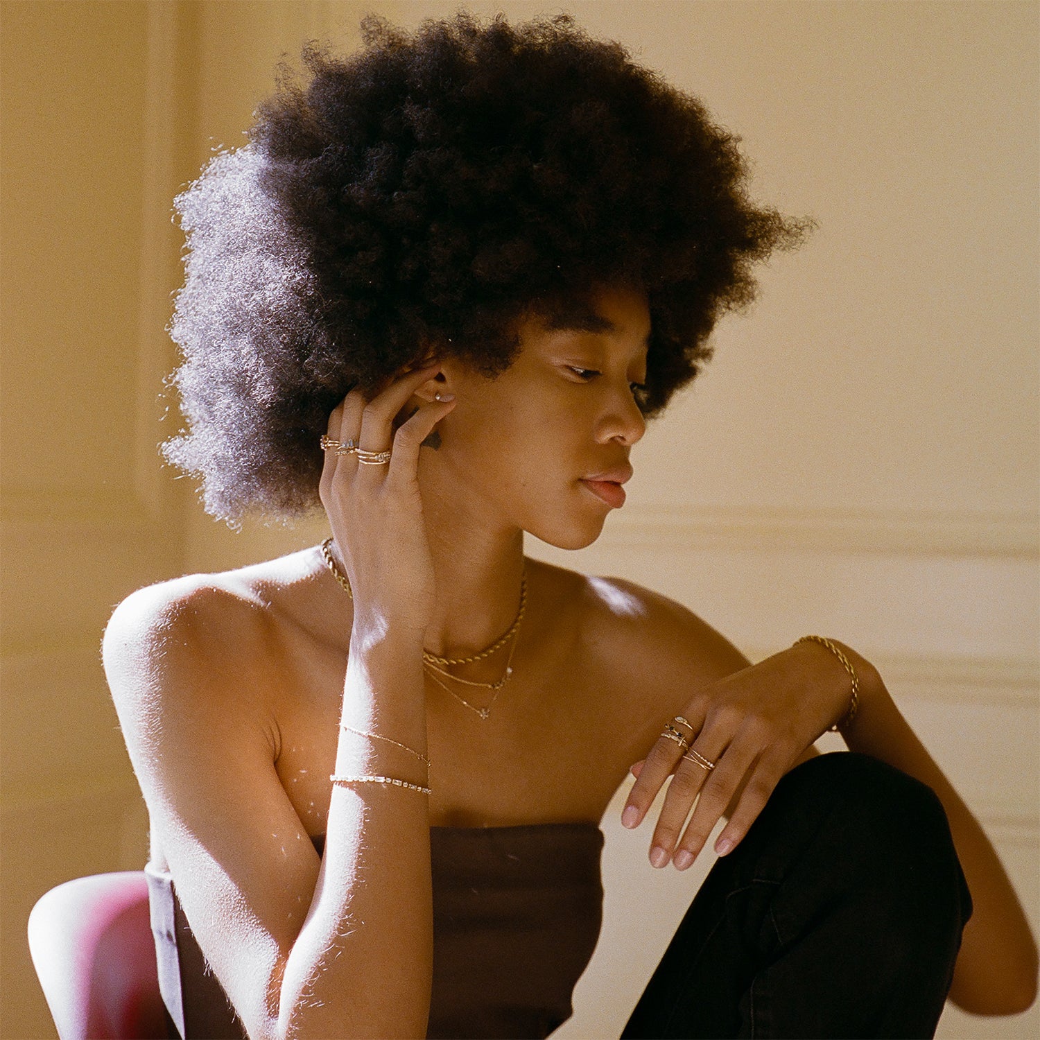 A woman with a natural afro sits in soft, warm light, gently touching her ear and looking down thoughtfully. She wears a strapless top and accessorizes with the Solid Gold Crossover Ring Set and other yellow gold jewelry.