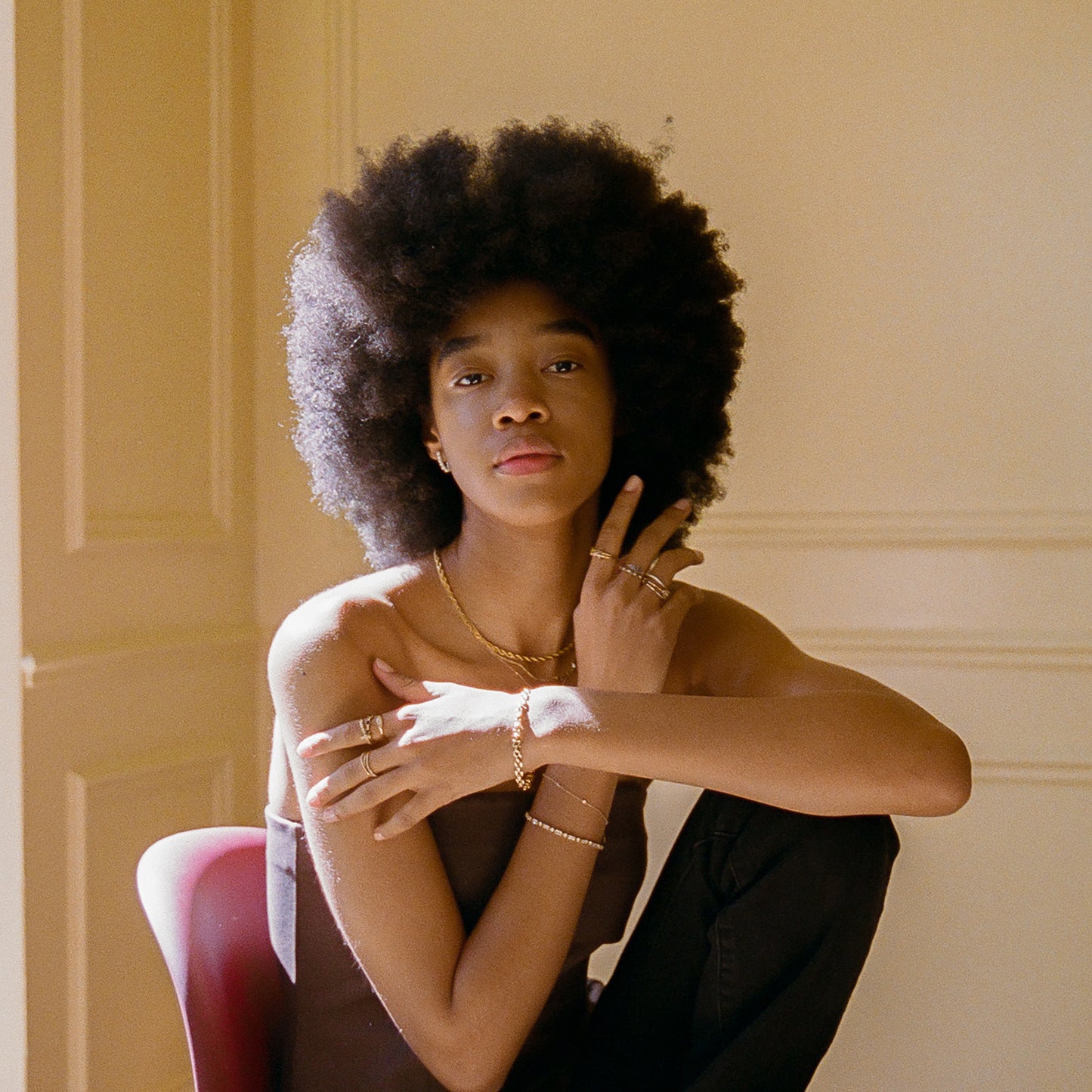 A person with a natural afro sits confidently on a red chair, wearing dark clothes and a Solid Gold Everyday Opal Bracelet. One arm rests on their knee as warm sunlight softly fills the room.