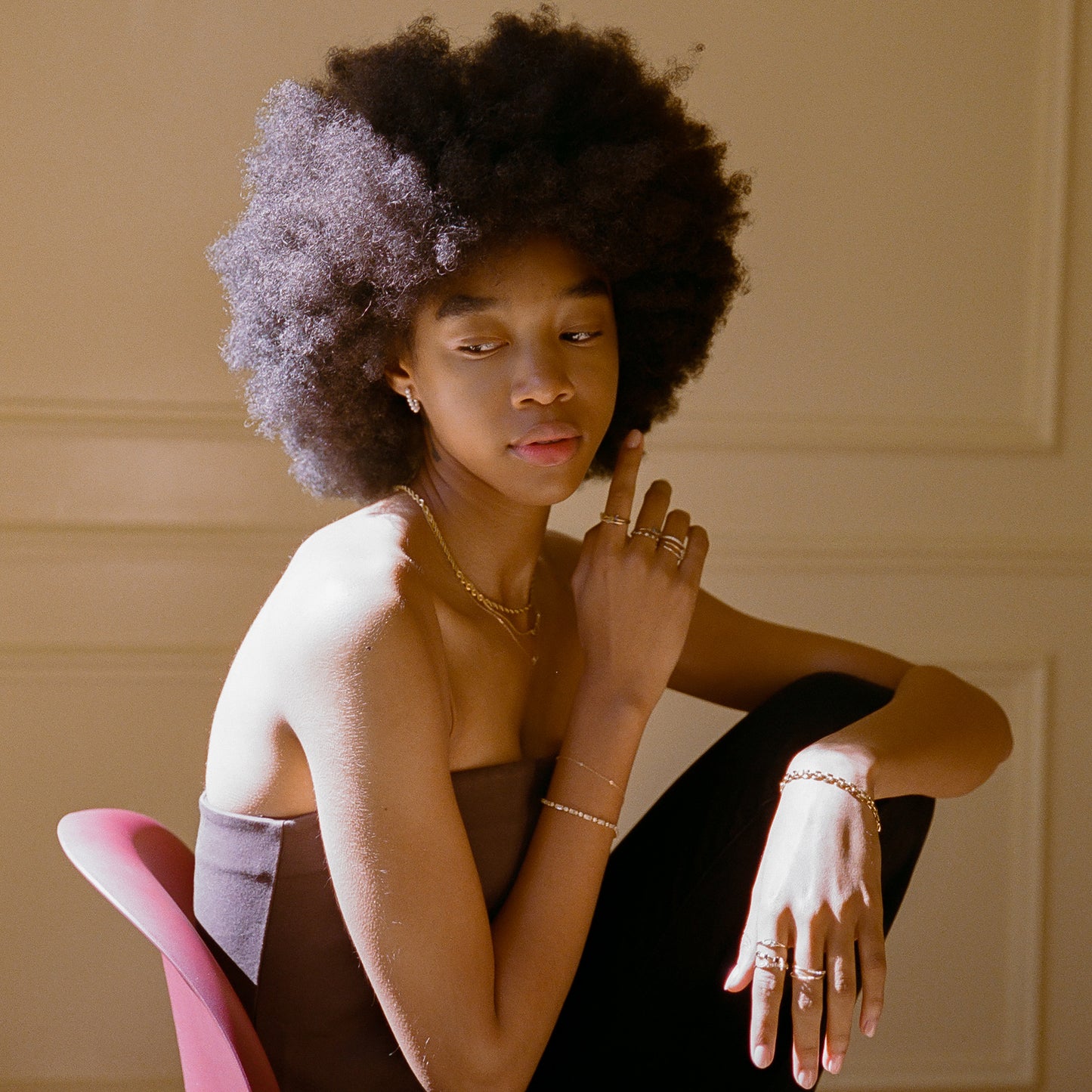 A woman with a natural afro sits sideways on a red chair in soft, warm light, wearing the Solid Gold Tourmaline Trio Stacking Ring and a strapless dark top, resting her chin on her hand and gazing thoughtfully to the side.