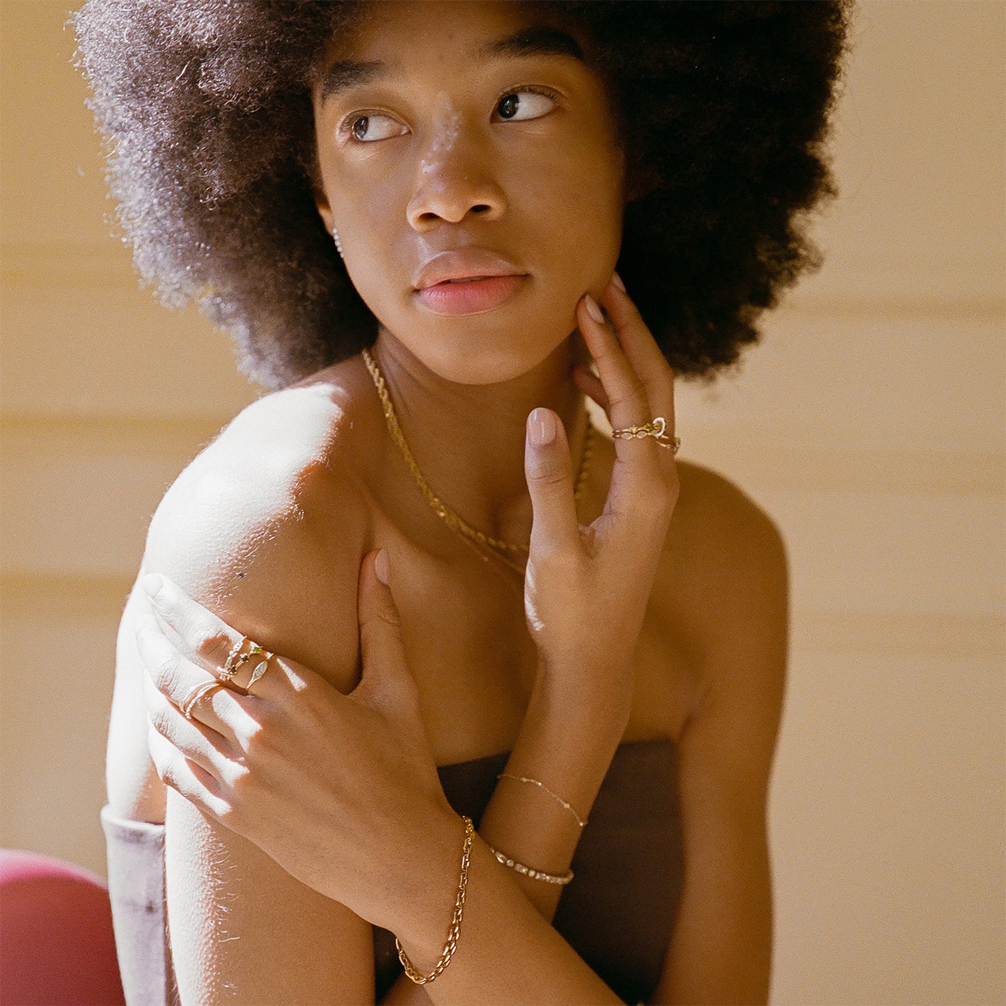 A woman with a natural afro gazes thoughtfully to the side, gently touching her neck and shoulder. She wears gold jewelry, including a Solid Gold Floating Gems Stacking Ring, bracelets, a necklace, and a strapless top.