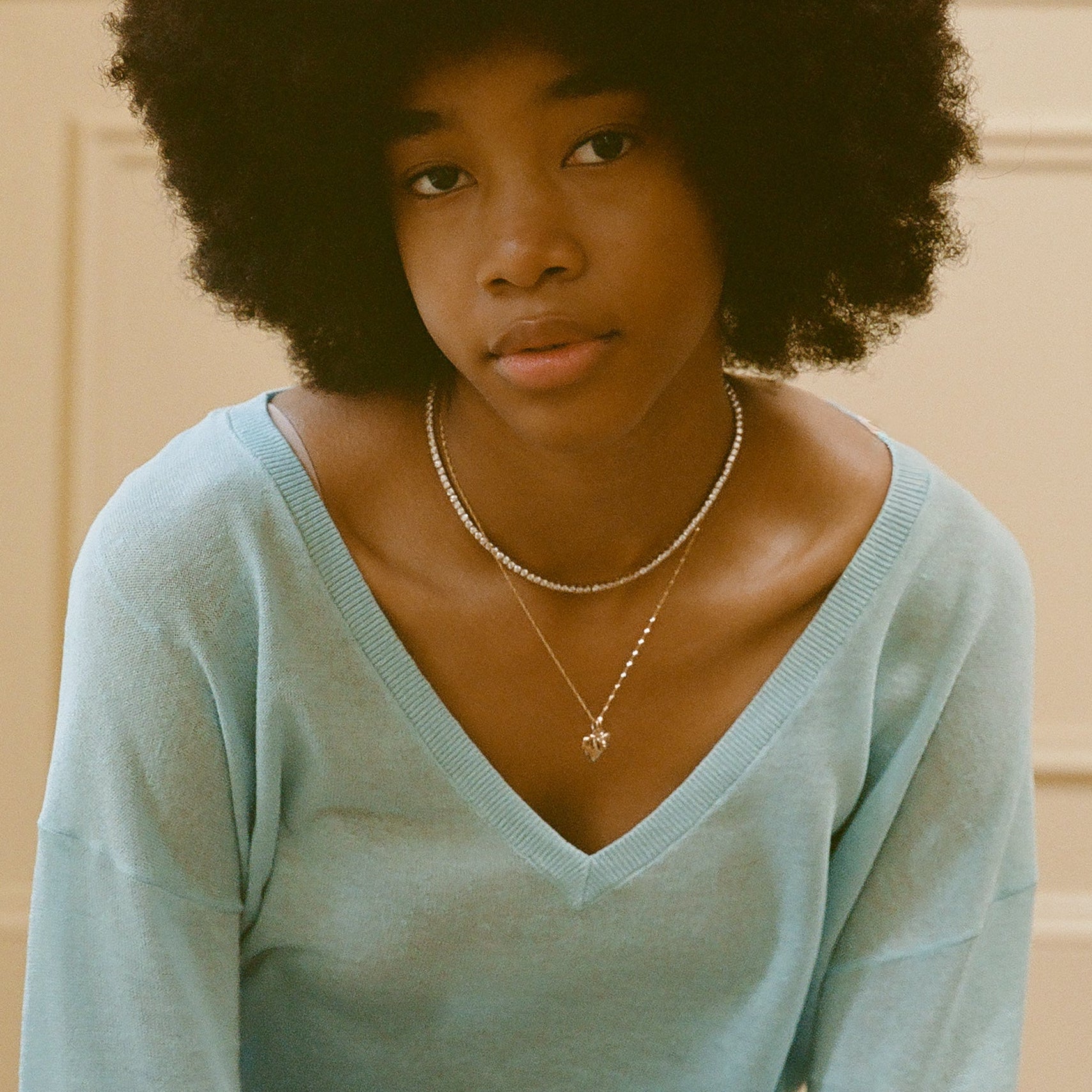 A young woman with a natural afro hairstyle wears a light blue V-neck sweater and two delicate necklaces, including the Tennis Necklace, and gazes softly at the camera against a neutral background.