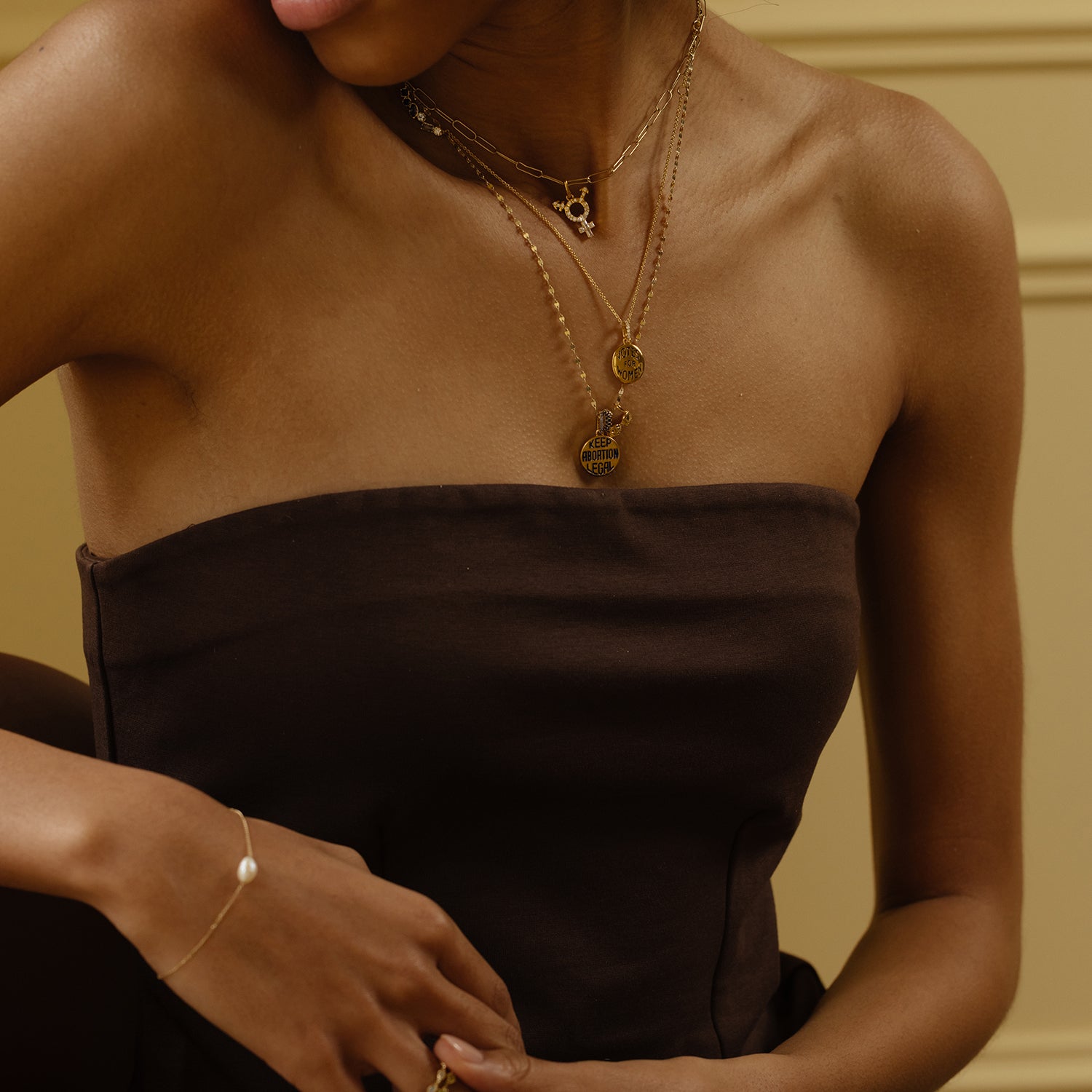 A woman in a strapless dark brown dress wears layered gold jewelry—including the Limited Edition Women's History Month Charm—while sitting against a beige background, celebrating unique women's charms.