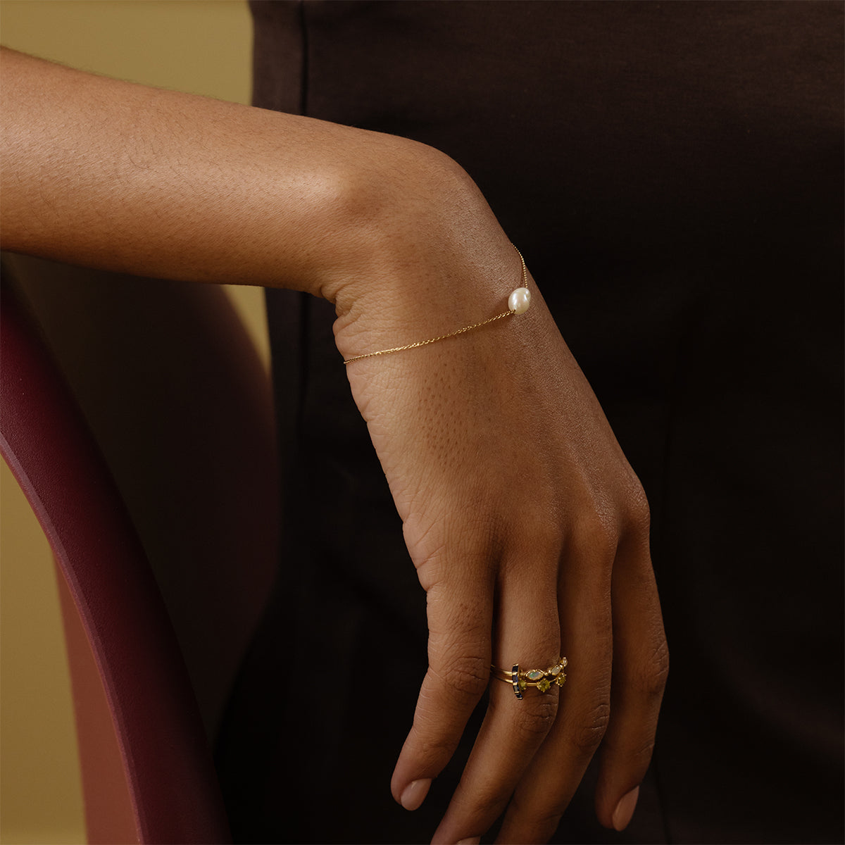A close-up of a hand resting on a maroon chair, wearing the Solid Gold Single Pearl Chain Bracelet and several gold rings with small stones. The person is dressed in a dark brown outfit.