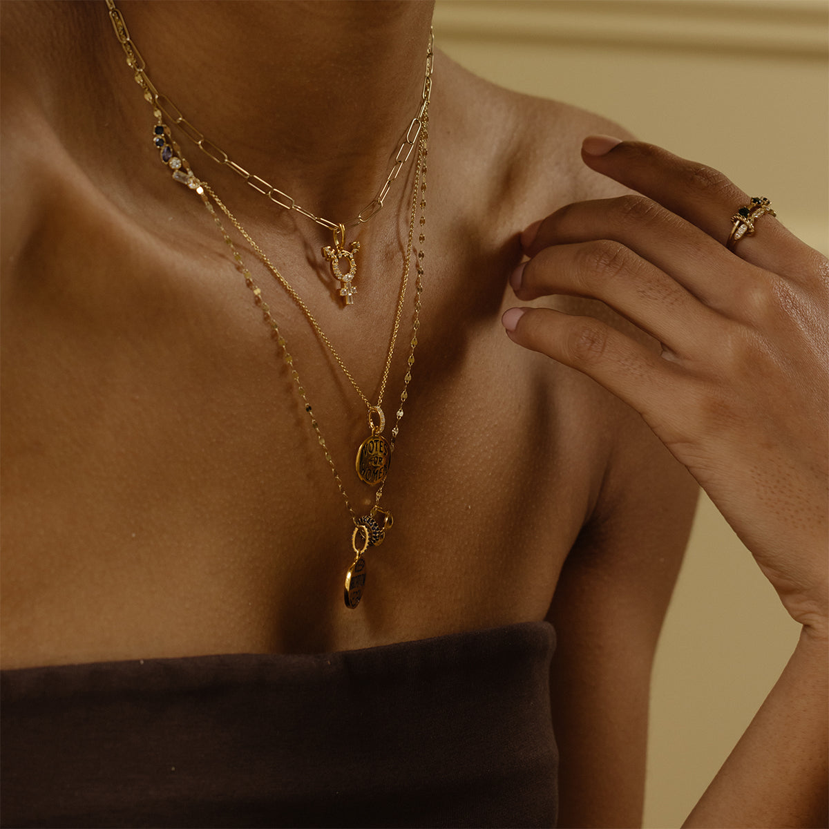 A person in a brown strapless top showcases layered gold necklaces with the Limited Edition Women's History Month Charm and a gold ring, set against a neutral background.