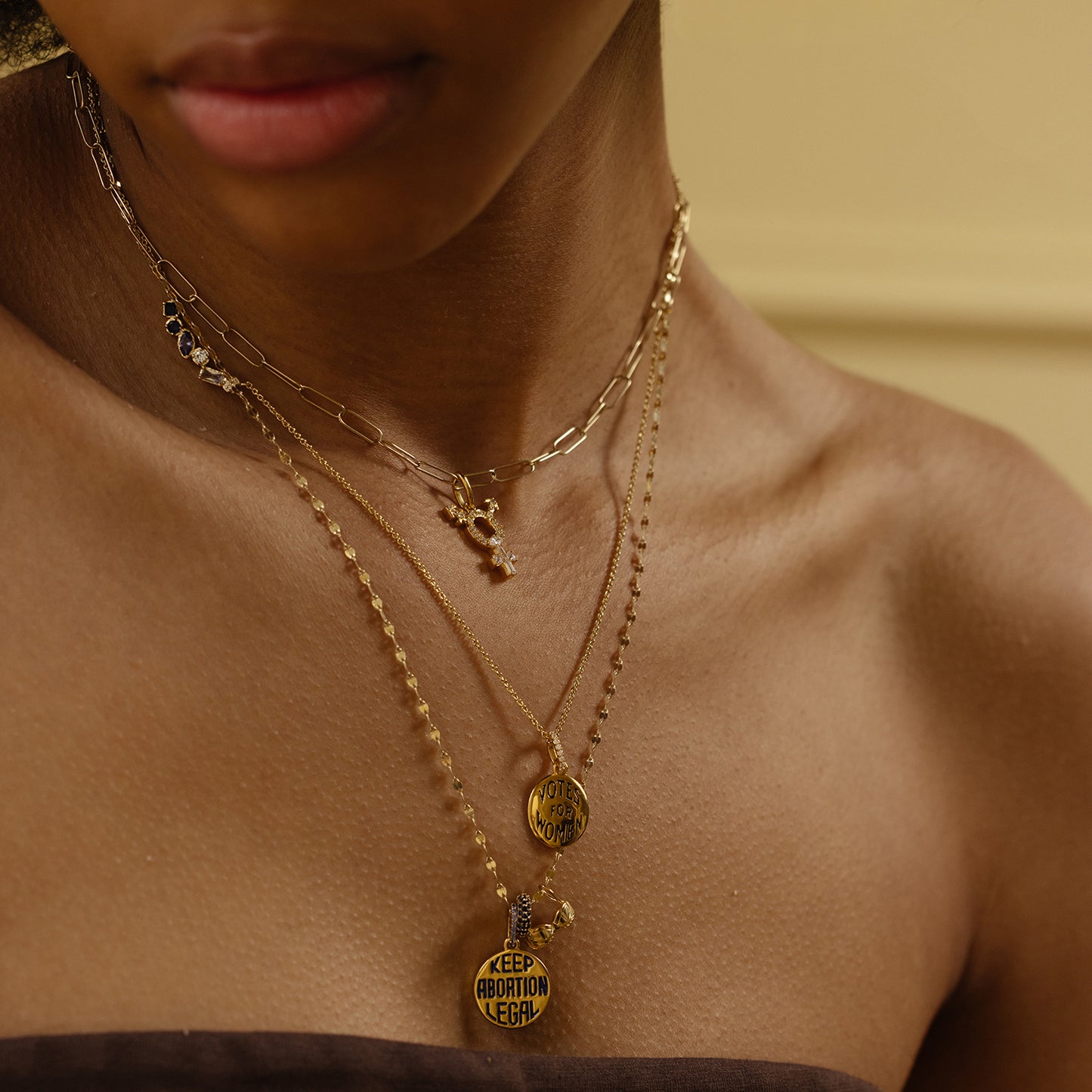 A close-up shows a person wearing three layered gold necklaces with Limited Edition Women's History Month Charms—one pendant reads KEEP ABORTION LEGAL and another says VOTE. Their neck, shoulders, and brown top are visible.
