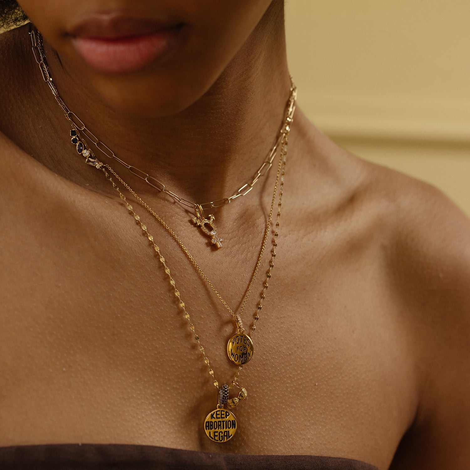 A close-up shows a person wearing three layered gold necklaces with Limited Edition Women's History Month Charms—one pendant reads KEEP ABORTION LEGAL and another says VOTE. Their neck, shoulders, and brown top are visible.