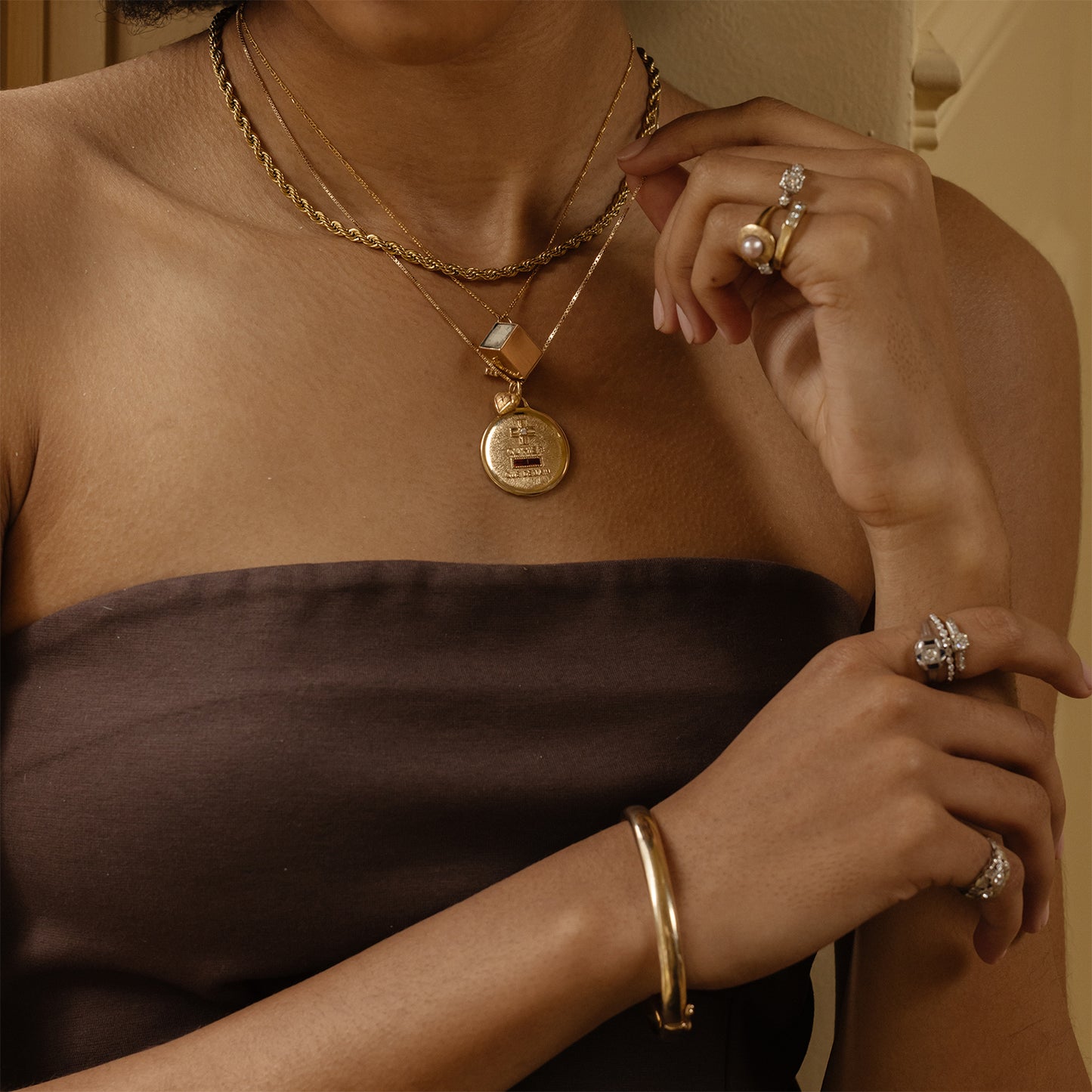 A woman in a strapless brown top shows off layered gold necklaces, gold rings with pearls and gemstones, and the Vintage 9k Classic Bangle, with her hands raised near her chest.