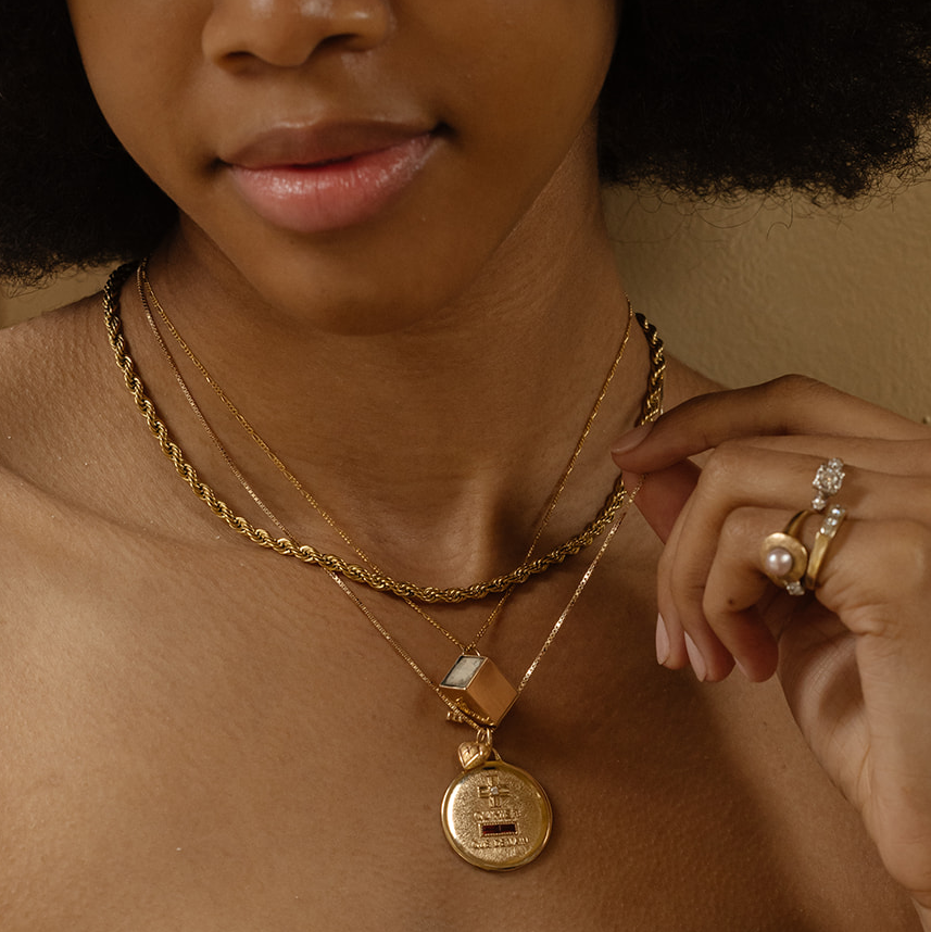A close-up of a woman wearing layered gold necklaces, including the 14k Vintage Revival Qu'hier Que Demain Pendant Charm. She also wears several gold rings, one adorned with a pearl and another with a diamond.
