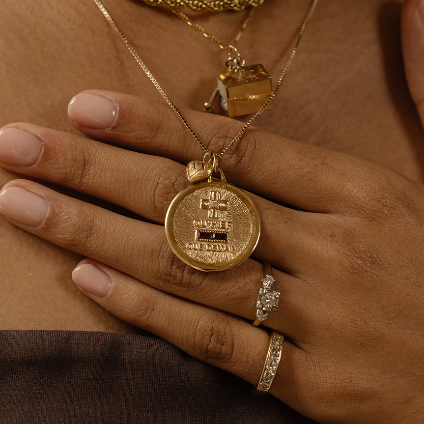 A close-up of a hand with nude nail polish and a diamond gold ring, resting on a chest adorned with layered gold necklaces, including the 14k Vintage Revival Qu'hier Que Demain Pendant Charm featuring a large, round, textured design.