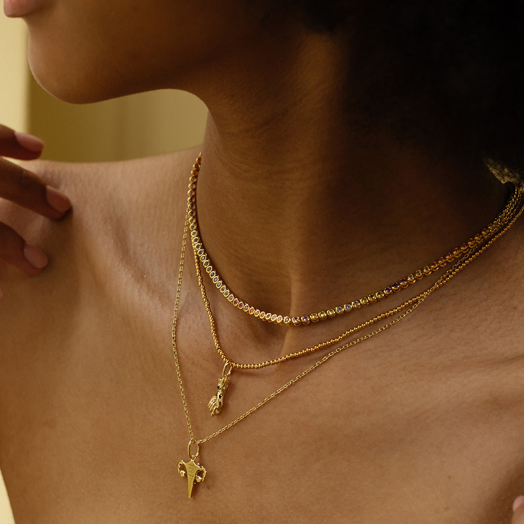 A person’s neck and collarbone, wearing three layered brass, 14k gold-plated necklaces with different pendants—including a CZ charm and small animal head—against a blurred background. Free Gift With Orders Over $200 - Choose A Charm.