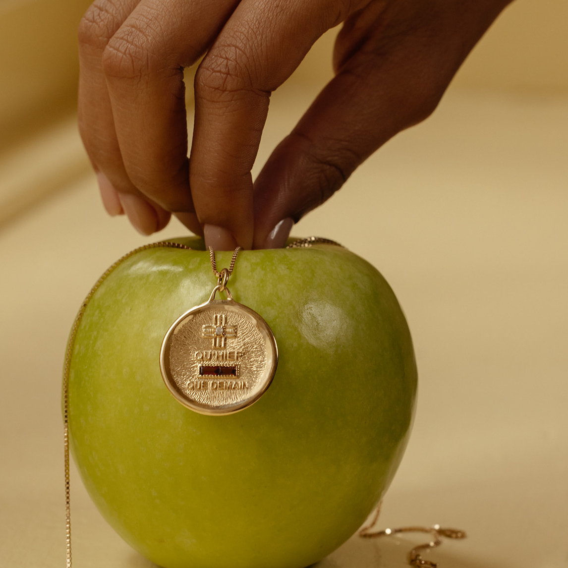 A hand holds the 14k Vintage Revival Qu'hier Que Demain Pendant Charm draped over a green apple, set against a soft beige background.
