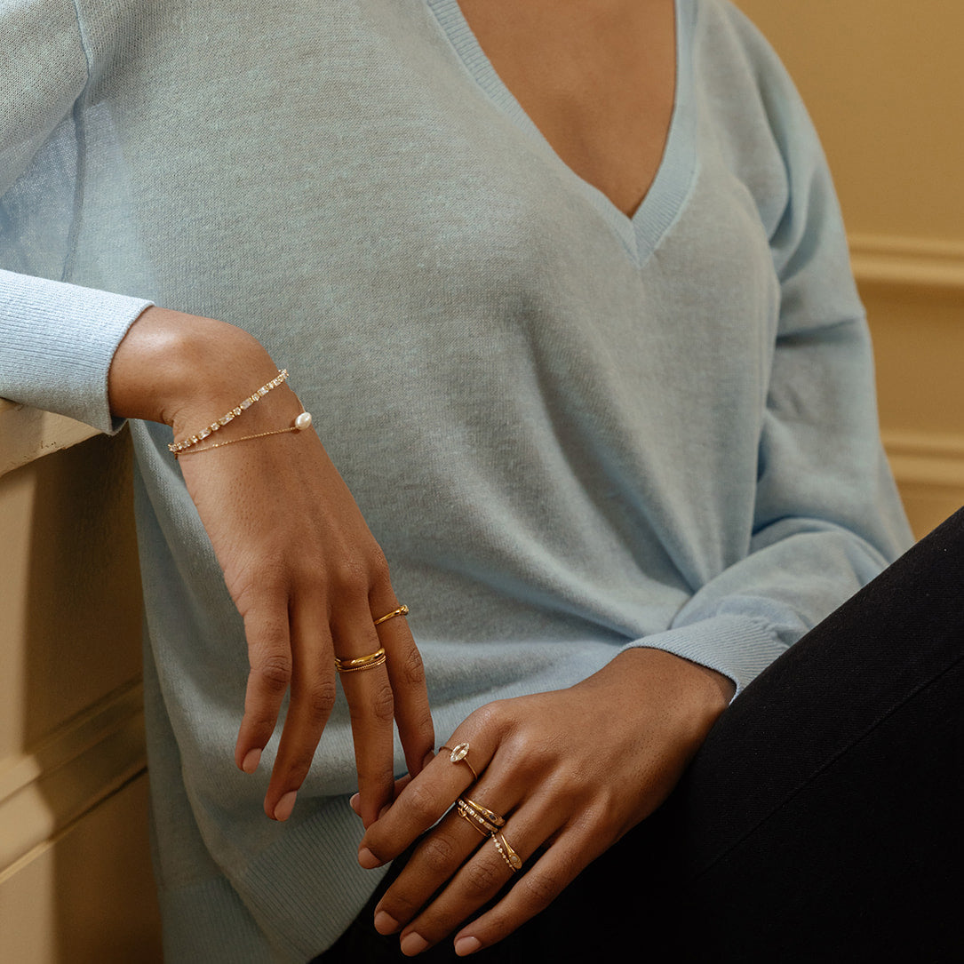 A person in a light blue V-neck sweater and black pants sits with arms resting on their knee, wearing multiple solid gold rings, including the Solid Gold Gemstone Marquise Ring in White Topaz, and a delicate gold charm bracelet.