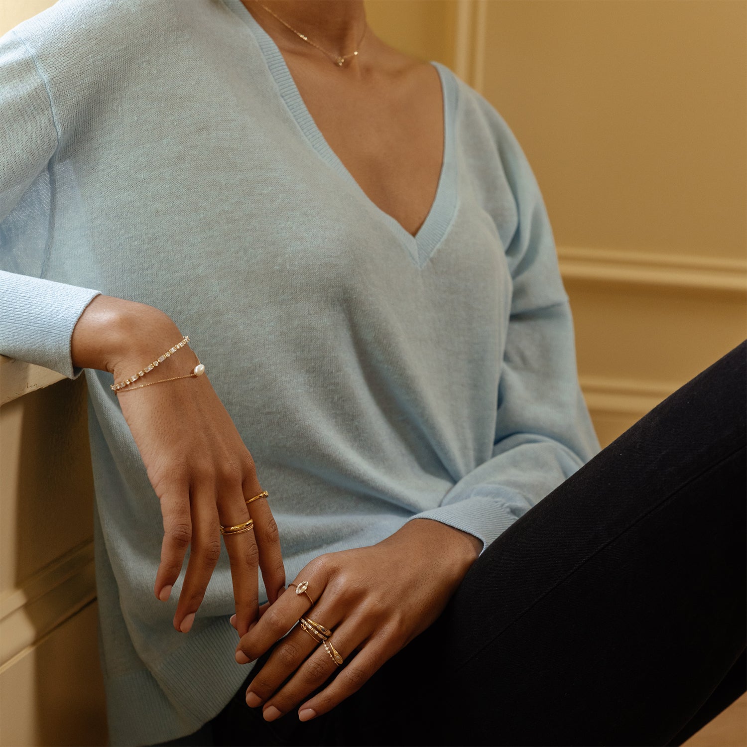 A woman indoors wears a light blue V-neck sweater, dark pants, several gold rings, a delicate necklace, and the Baguette Tennis Bracelet in white CZ with 14k yellow gold plating. Her face is partially obscured.