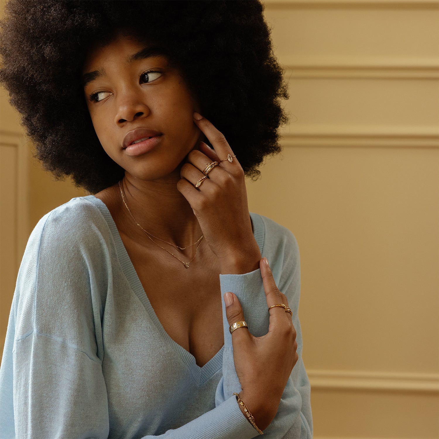 A woman with an afro and a light blue sweater wears the Solid Gold Pizza Acrostic Ring and gold necklaces, gazing thoughtfully to the side. She touches her face with one hand; a cream paneled wall is behind her.