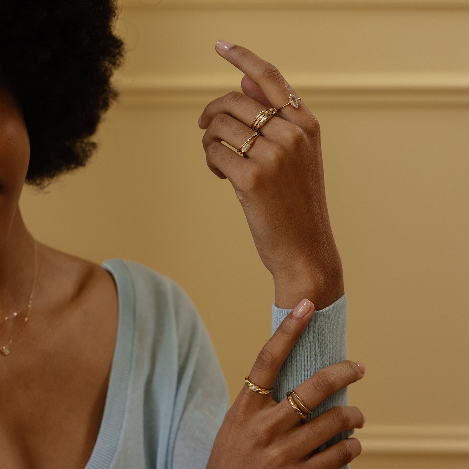 A person with medium brown skin and curly hair wears the Solid Gold Crossover Ring Set and a delicate necklace, showing their hands against a beige background while wearing a light blue top.